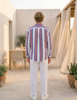 Man wearing a striped shirt and white pants standing in an outdoor setting with plants and a table.