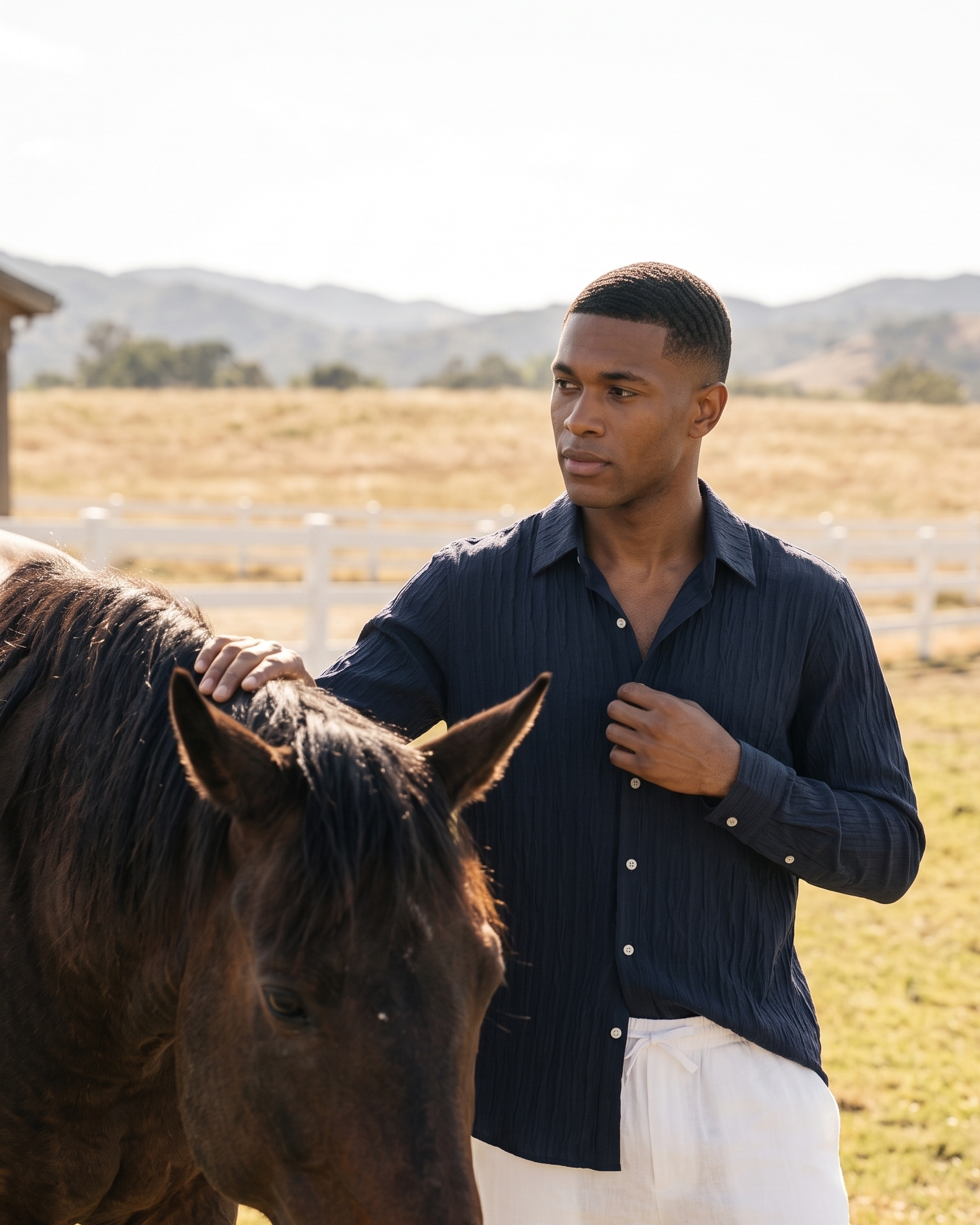 A man in a navy blue ribbed button-down shirt and white pants stands in a sunny outdoor paddock next to a dark brown horse, with a white fence and hills in the background.