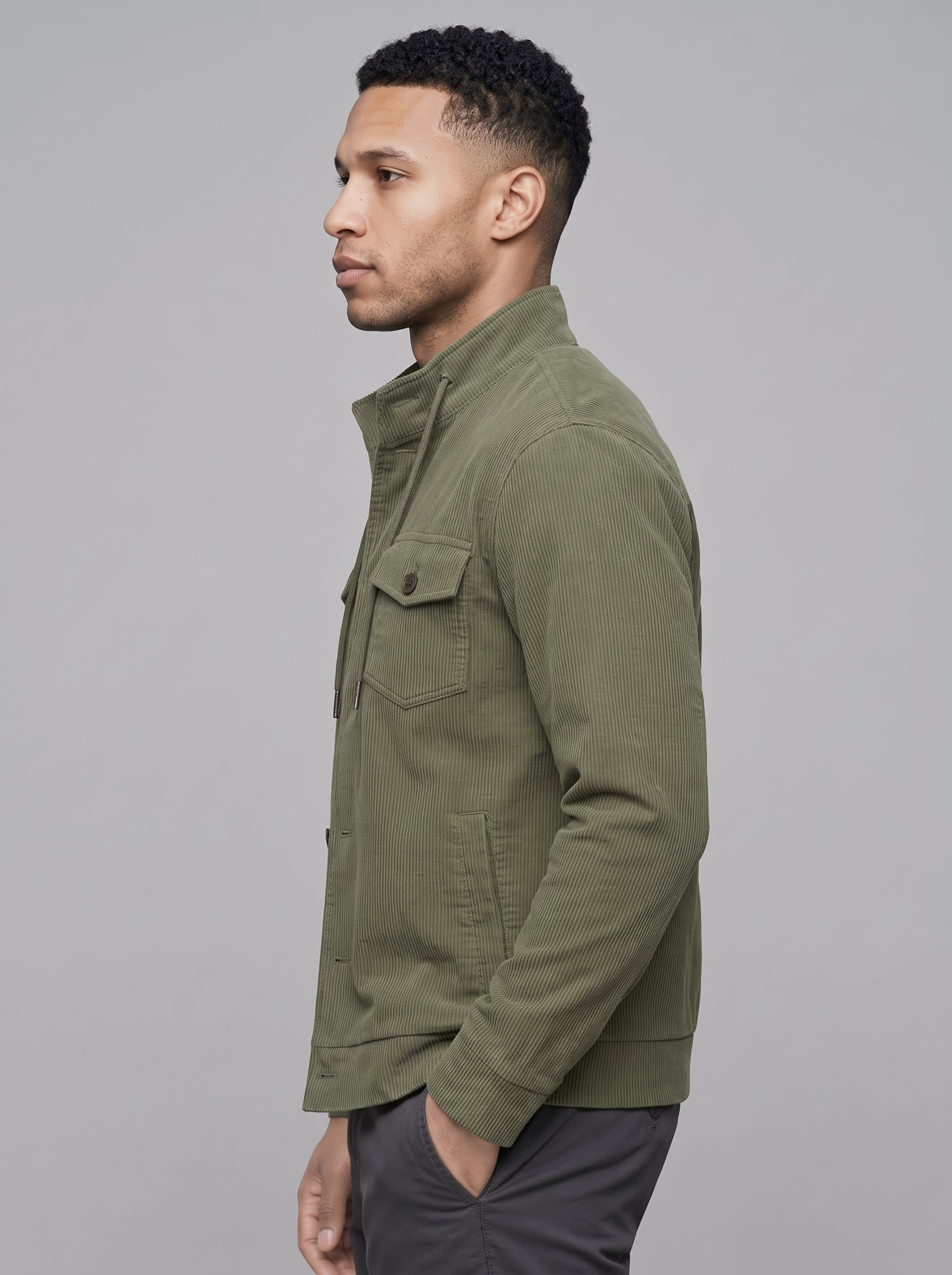A man in profile wearing an olive green utility jacket and dark gray trousers, photographed against a solid gray studio background.