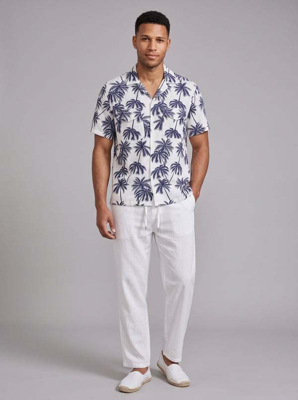 A man modeling a white short-sleeve button-up shirt with a navy blue palm tree pattern, paired with white drawstring trousers and white slip-on shoes, against a solid gray studio background.