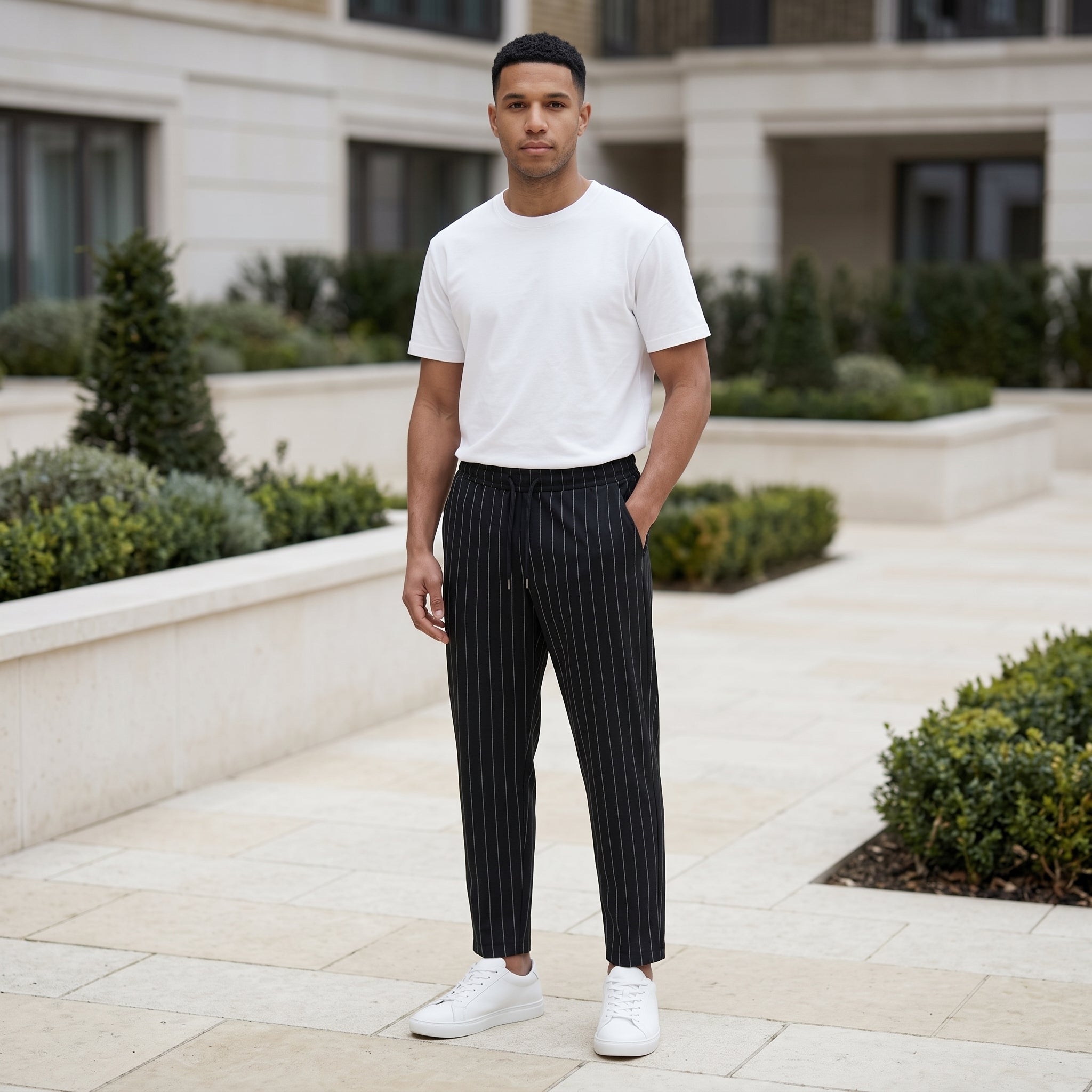 A man standing outdoors in a landscaped courtyard wearing a white crew-neck t-shirt, black pinstriped trousers, and white sneakers.