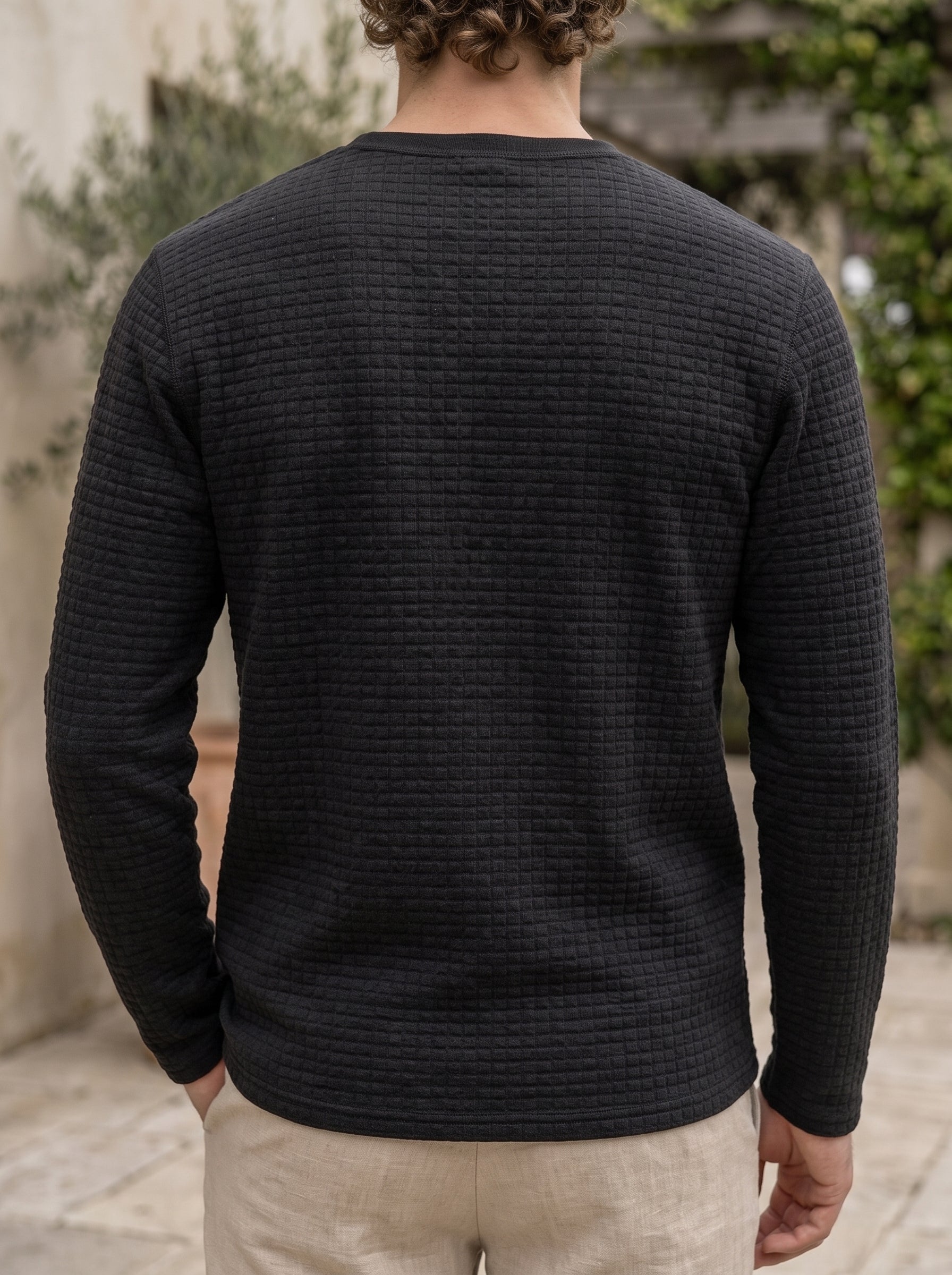 A man viewed from behind wearing a dark charcoal gray waffle-knit long-sleeve shirt and light beige trousers, standing outdoors against a blurred stone wall and greenery background.