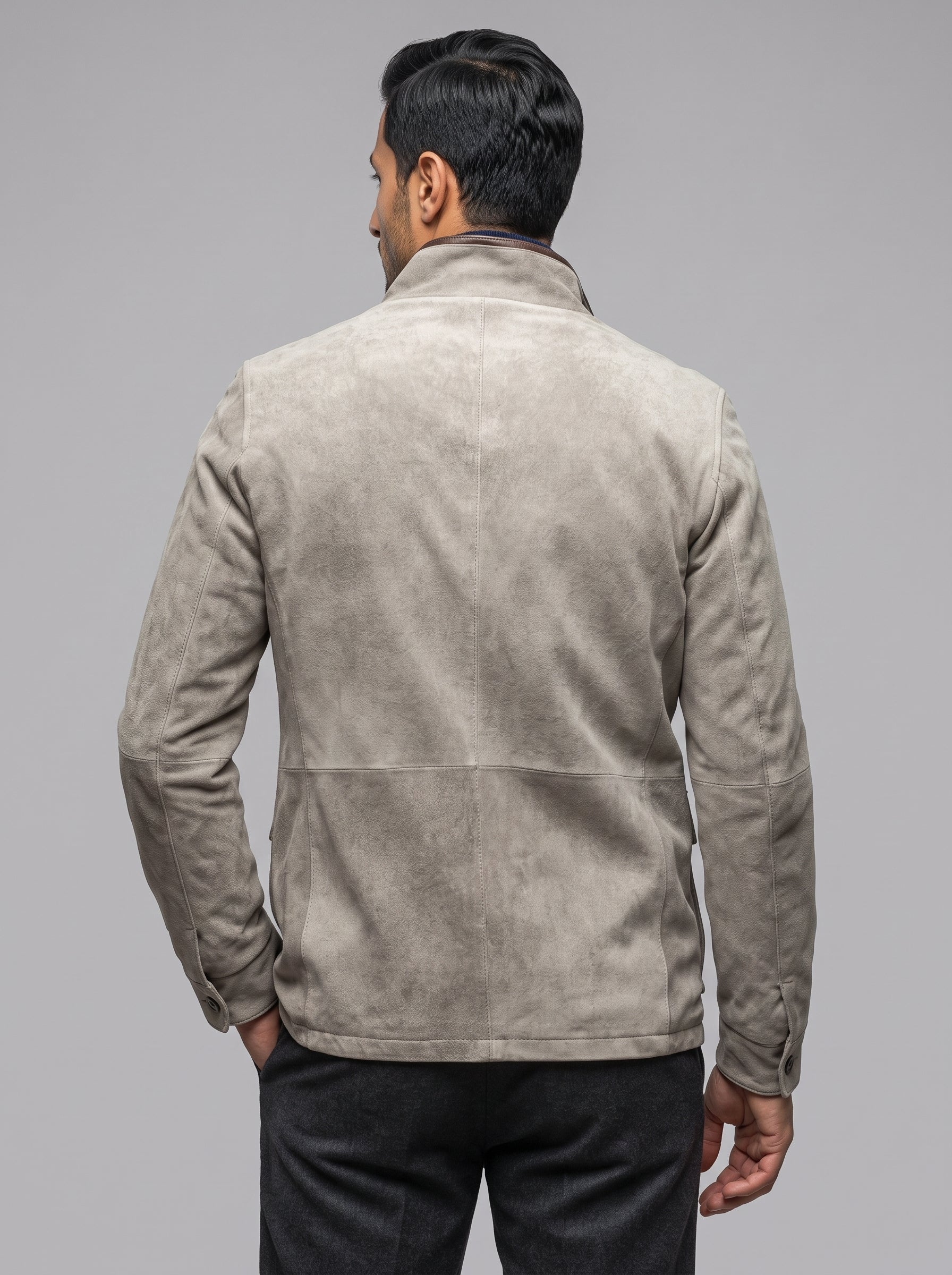 A man viewed from behind wearing a light taupe suede jacket and dark trousers, standing against a solid gray studio background.