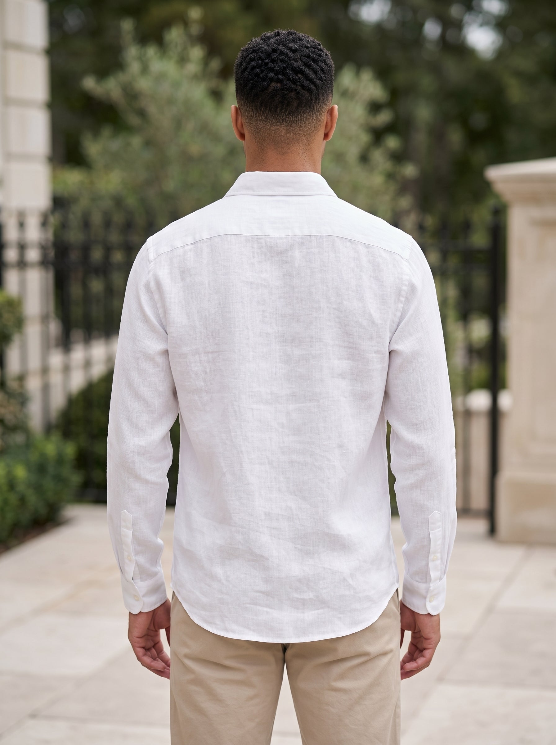 A man viewed from behind wearing a white long-sleeve linen button-down shirt and khaki pants, standing outdoors on a stone walkway.