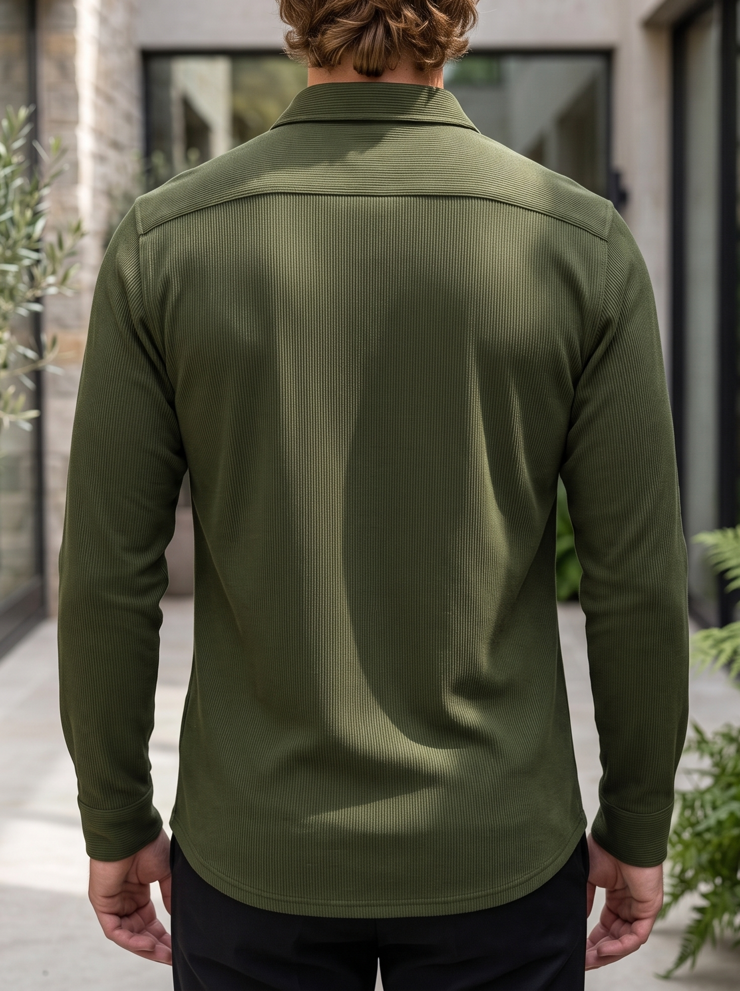 A man viewed from behind wearing an olive green long-sleeve button-up shirt, standing in an outdoor patio setting.