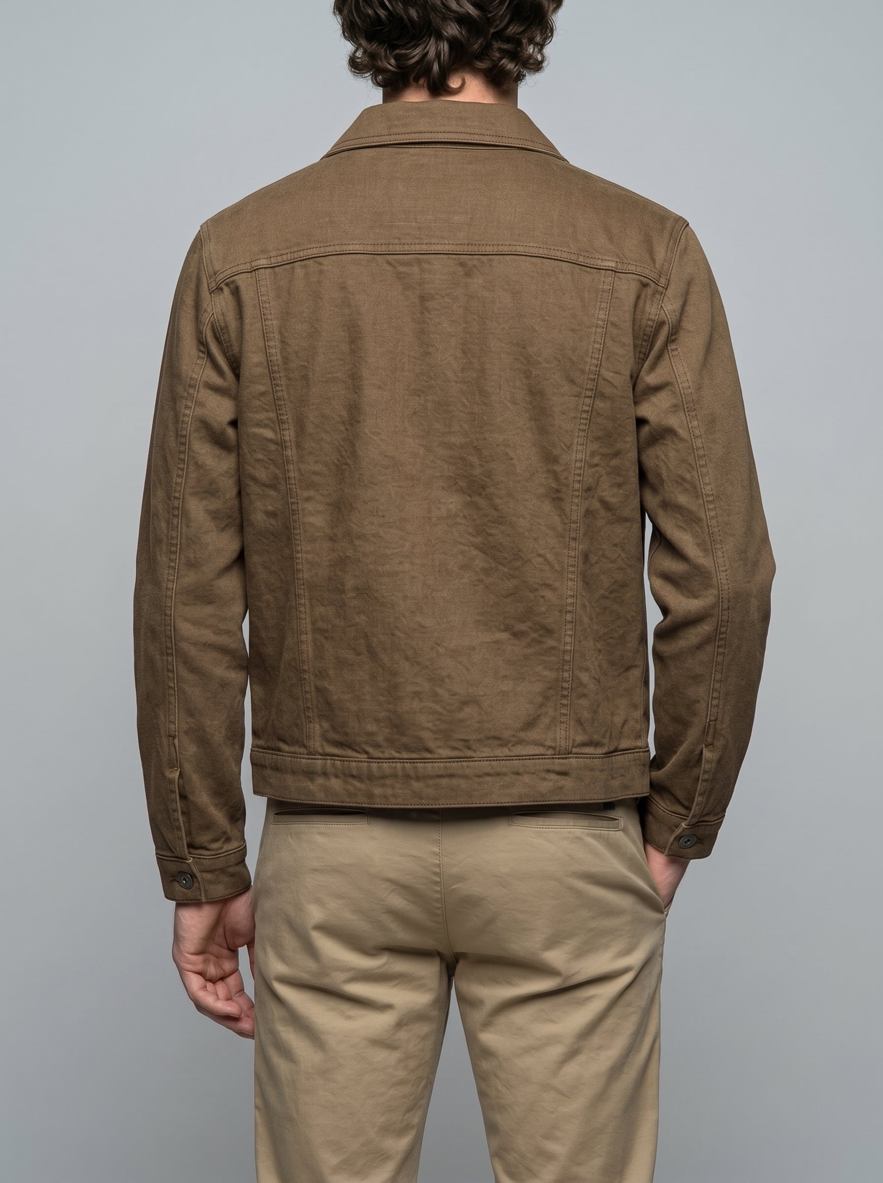 A man viewed from the back wearing a brown linen-textured jacket and beige trousers, set against a solid gray studio background.