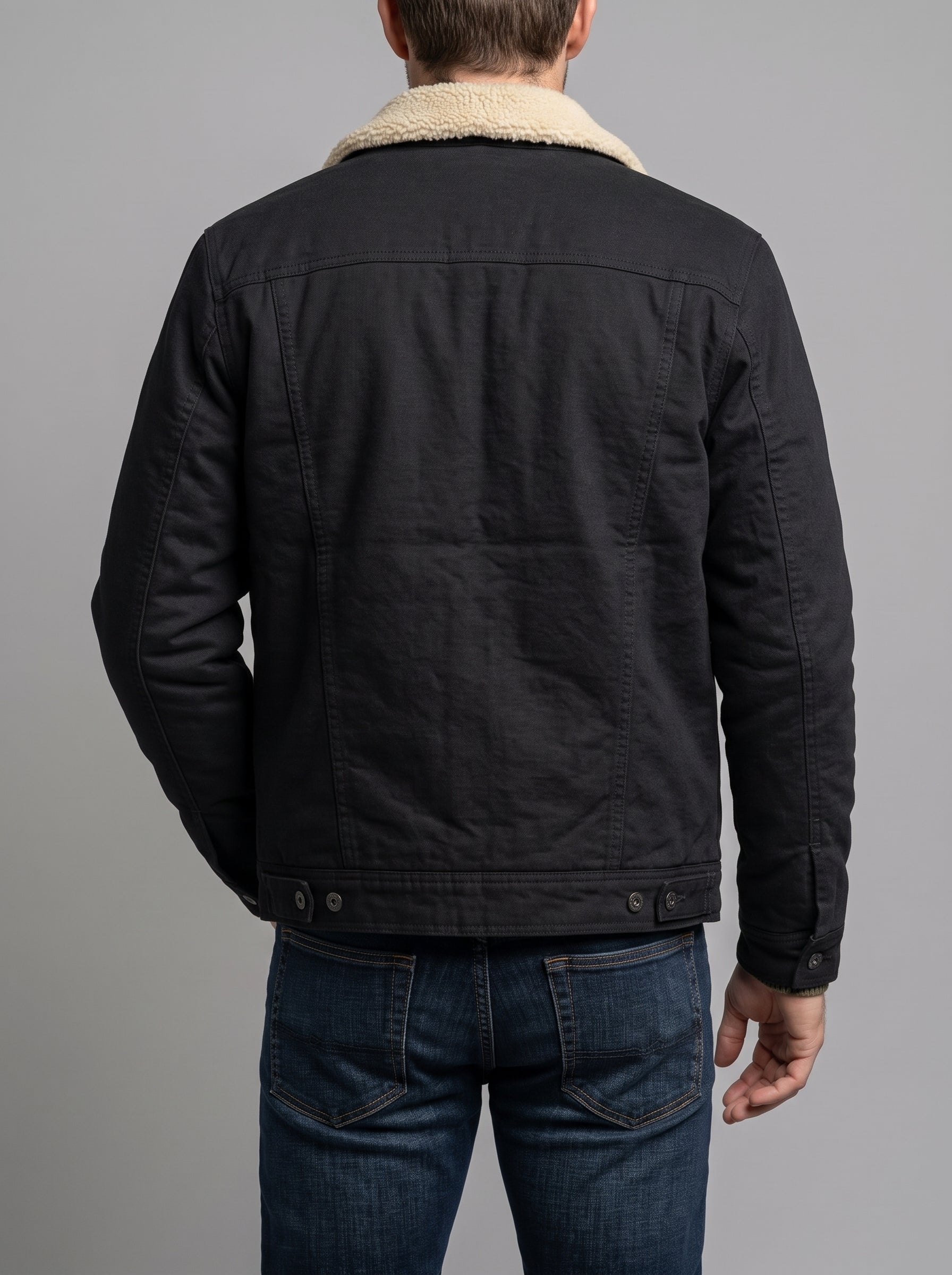 A man viewed from the back wearing a black quilted jacket with a beige sherpa collar and blue jeans, set against a solid gray studio background.