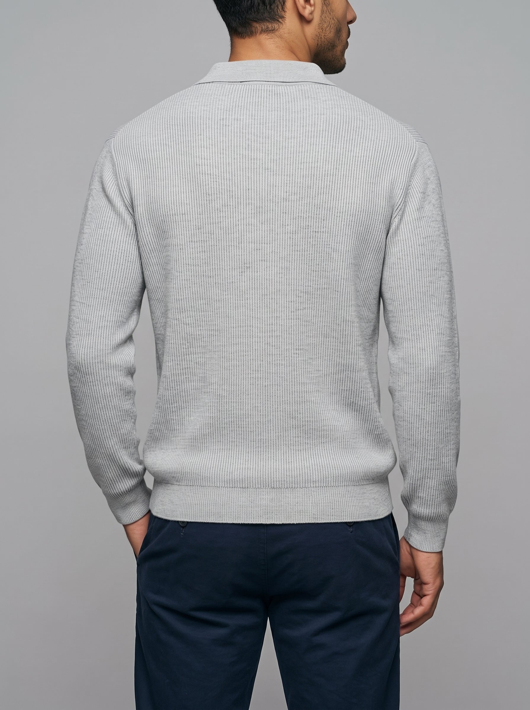 A man viewed from the back wearing a light gray ribbed knit sweater and navy blue trousers, set against a solid gray studio background.