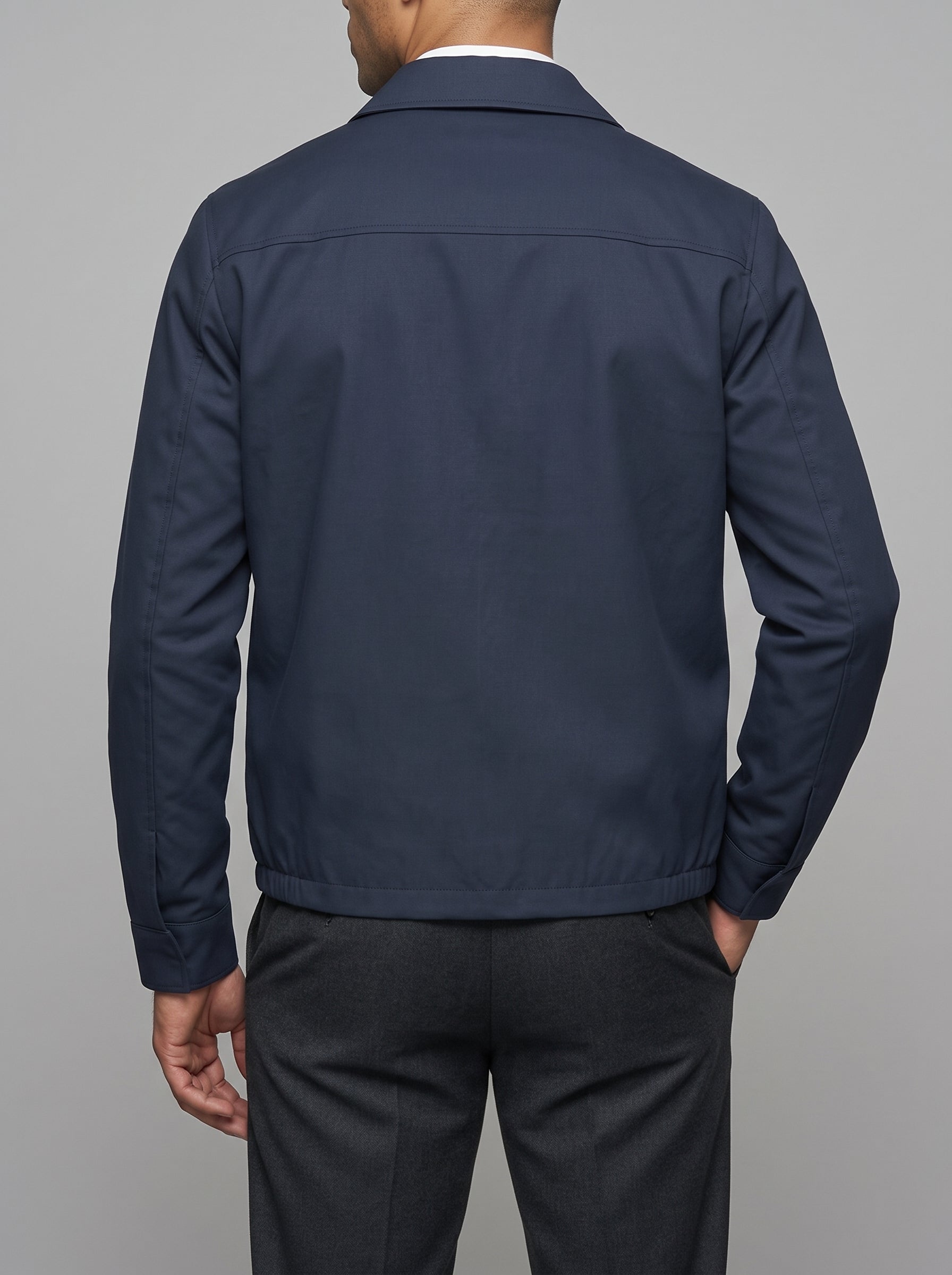 A man viewed from the back wearing a navy blue jacket over a white collared shirt and dark gray trousers, set against a solid gray studio background.