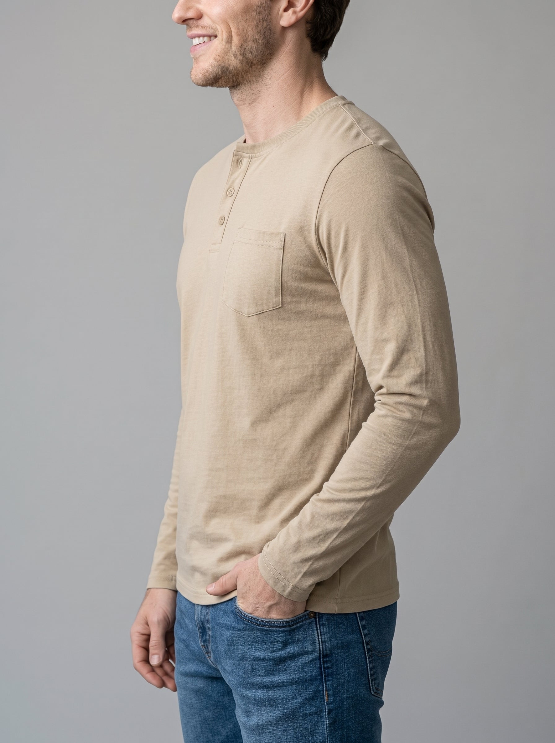 A man wearing a beige long-sleeve henley shirt with a chest pocket, paired with blue jeans, shown in a side profile against a solid gray background.