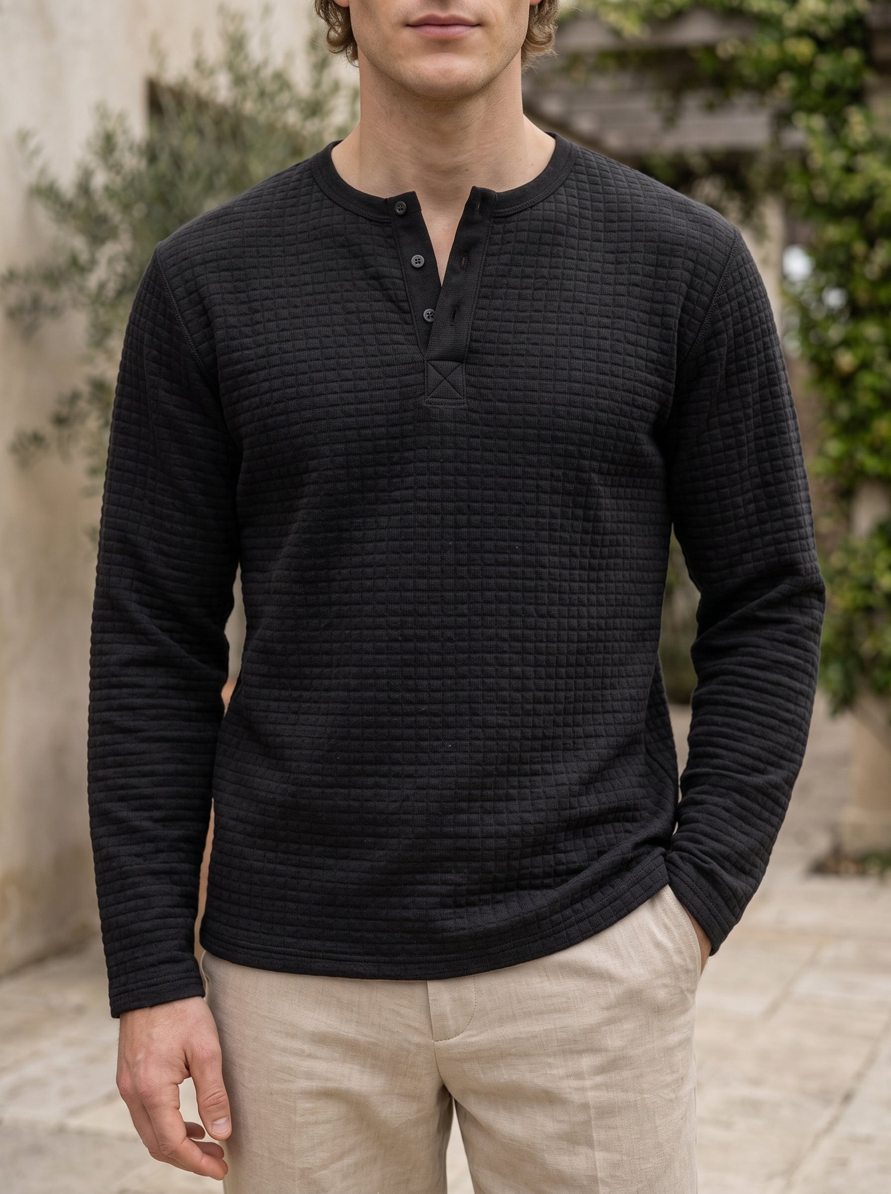 A man wearing a black long-sleeve waffle-knit henley shirt with a three-button placket, paired with beige trousers, standing outdoors against a blurred stone wall and greenery background.