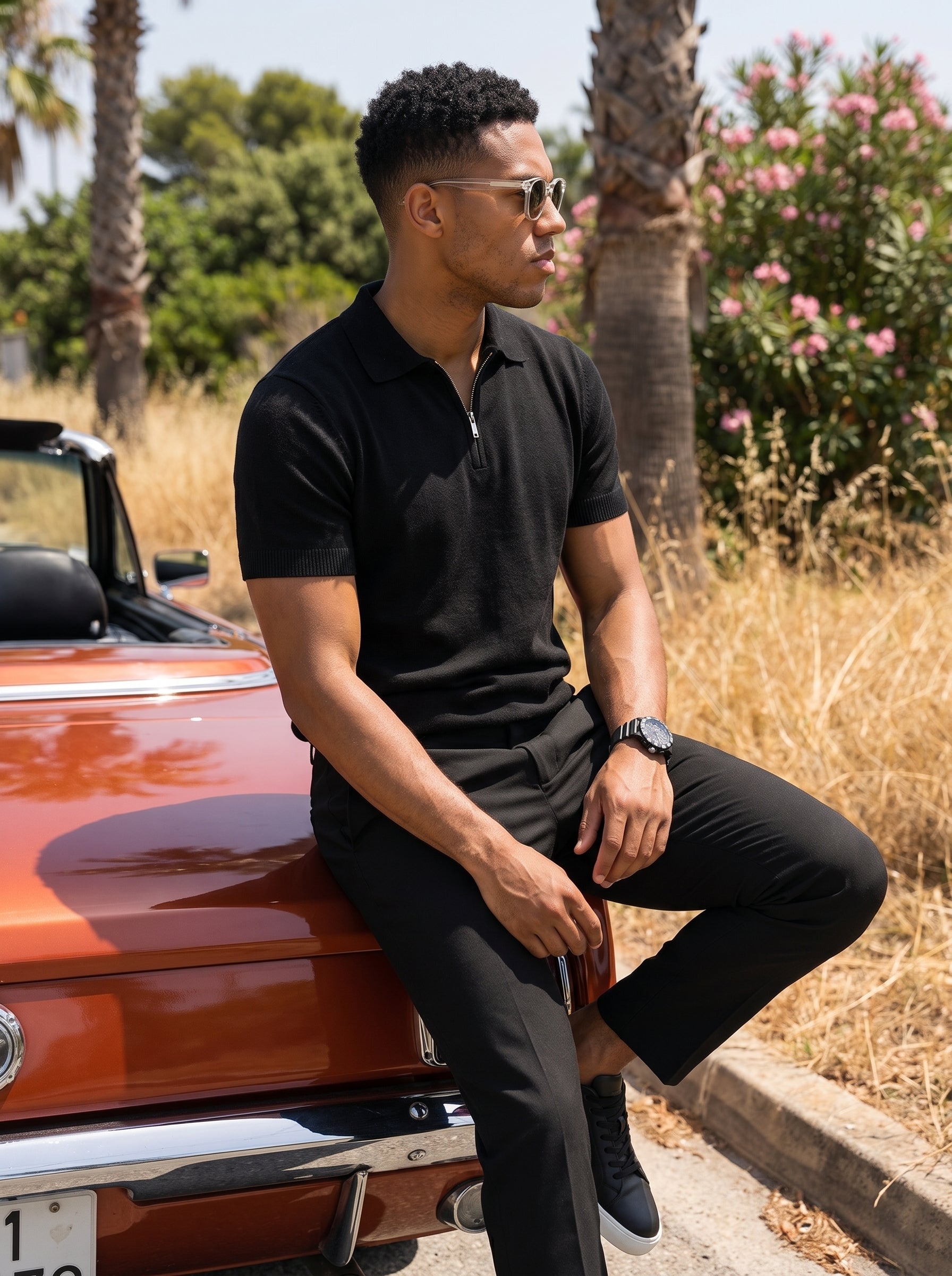 A man wearing a black short-sleeve polo shirt and black pants sits on the front bumper of a rusted orange classic convertible car outdoors, with palm trees and dry grass in the background.