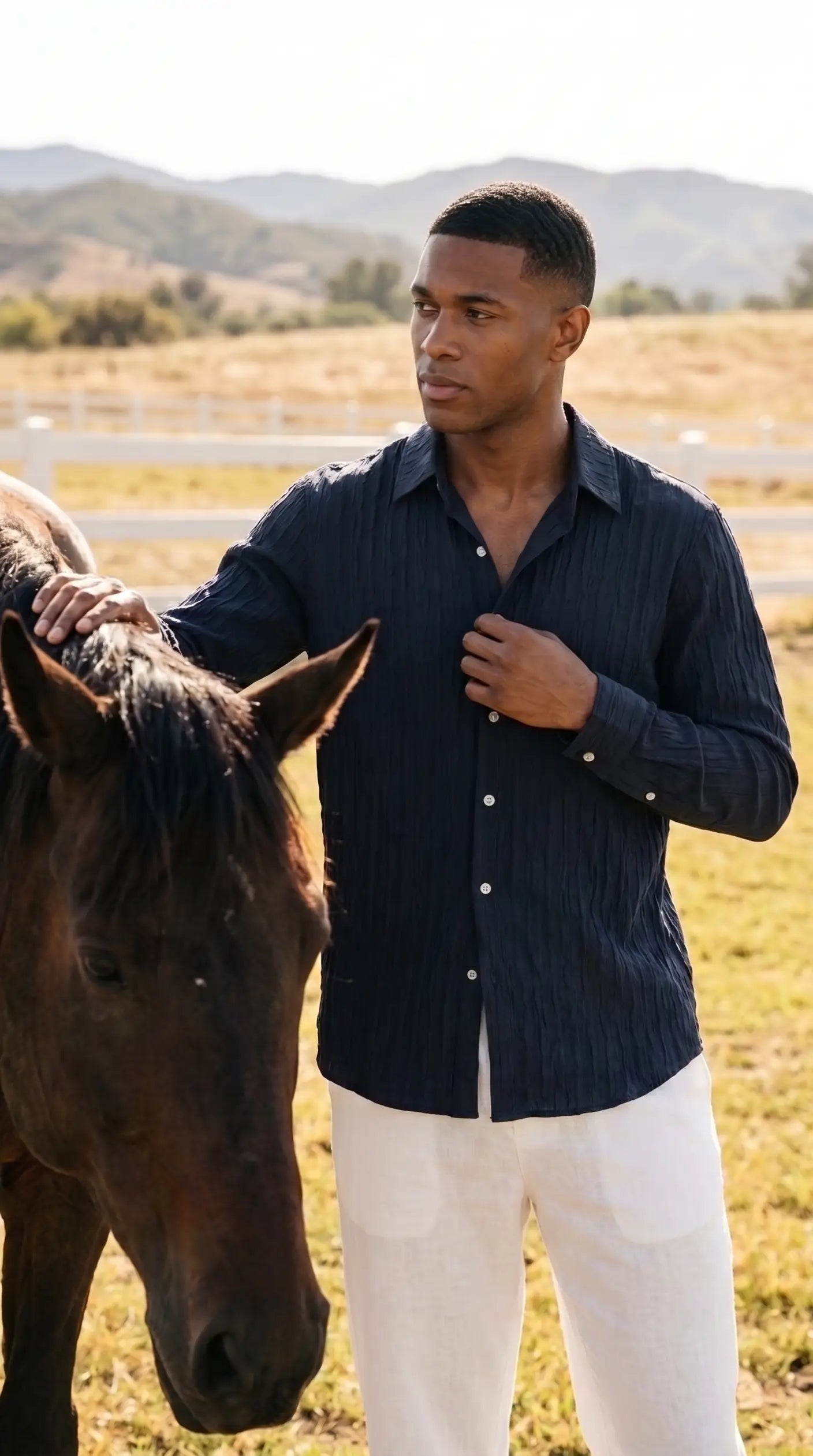 A man wearing a dark navy blue vertically striped button-down shirt and white pants stands in a sunny outdoor field next to a brown horse.
