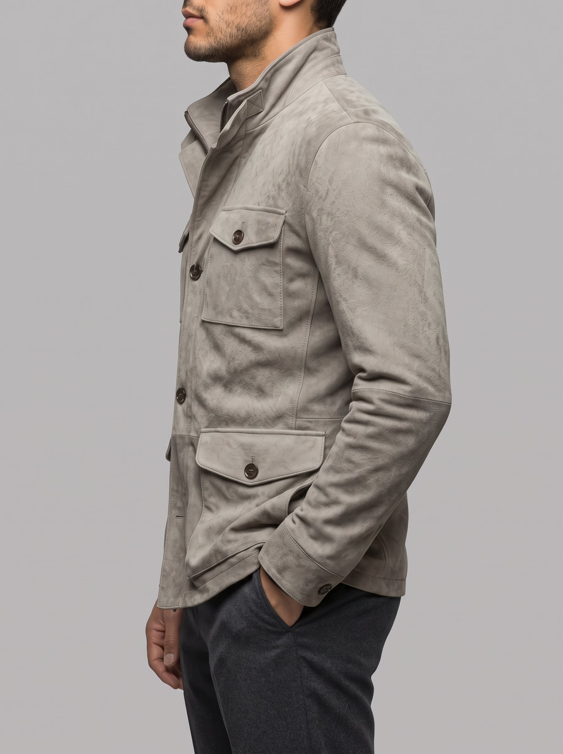 A man wearing a light gray suede jacket with dark buttons and flap pockets, paired with dark gray trousers, shown in a side profile against a solid gray background.