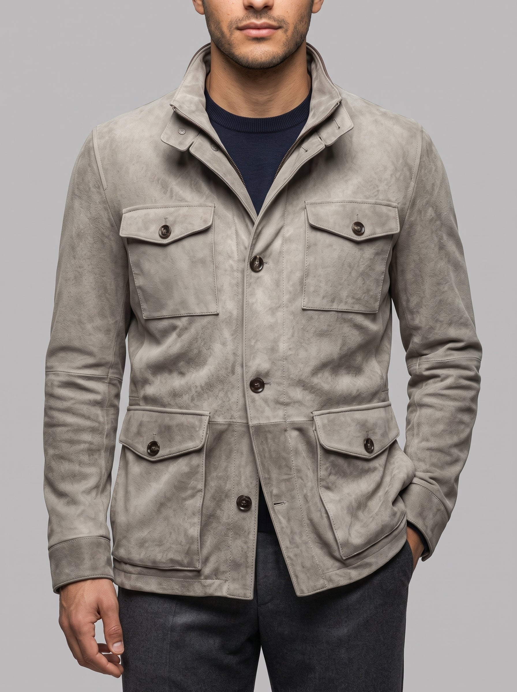 A man wearing a light gray suede utility jacket over a navy blue t-shirt and dark gray jeans, photographed against a solid gray studio background.