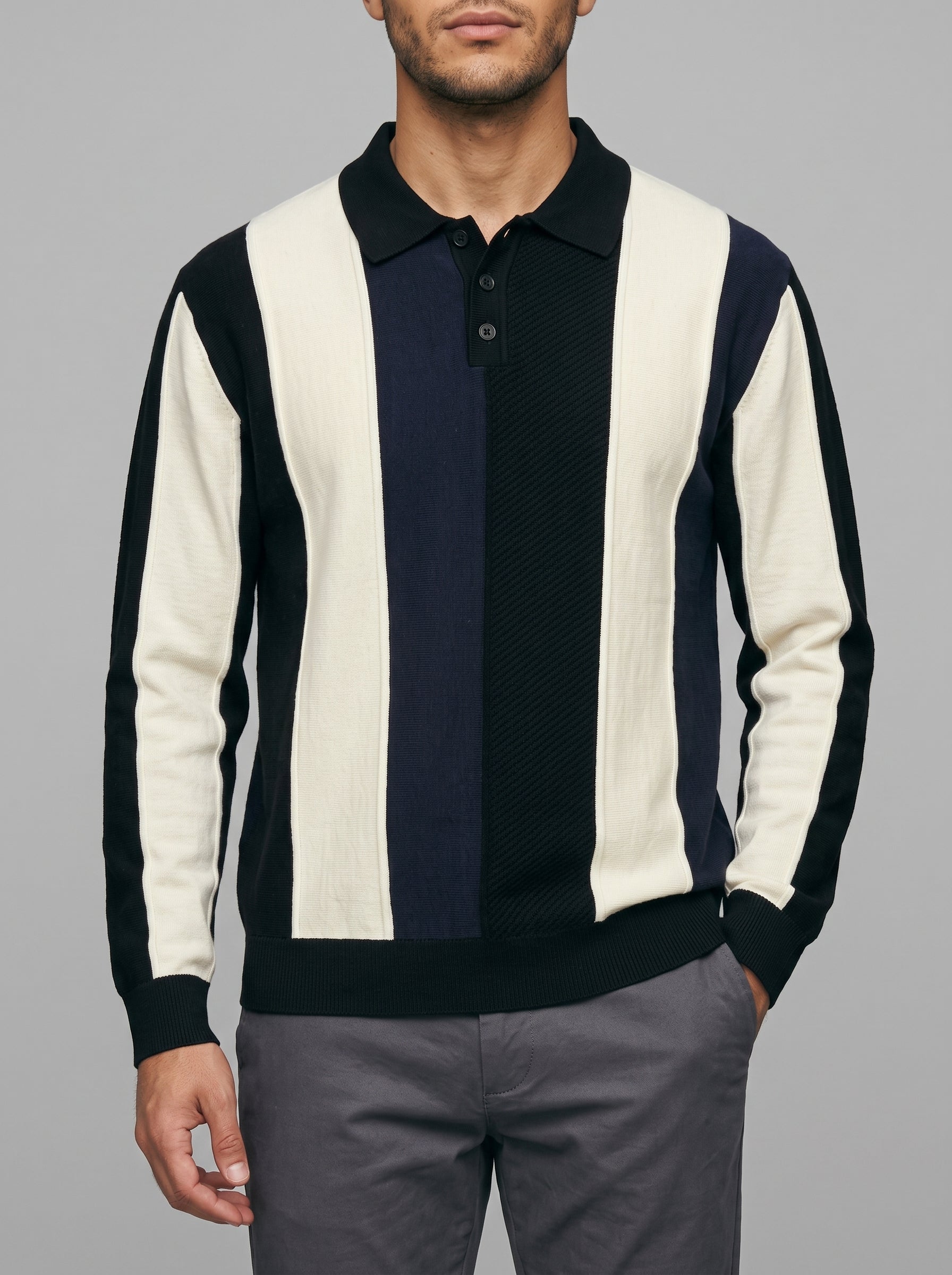 A man wearing a long-sleeve knit polo sweater with vertical stripes in cream, navy, and black, paired with gray trousers, against a solid gray studio background.
