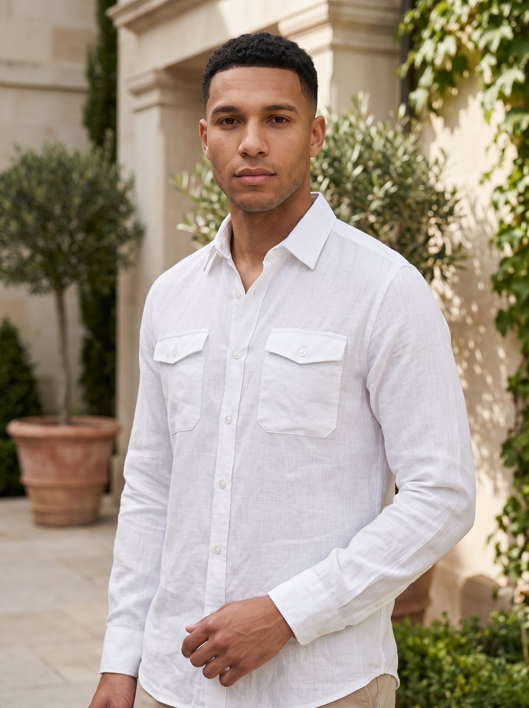 A man wearing a white long-sleeve linen button-up shirt with two chest pockets, standing outdoors in front of a beige building with green plants.