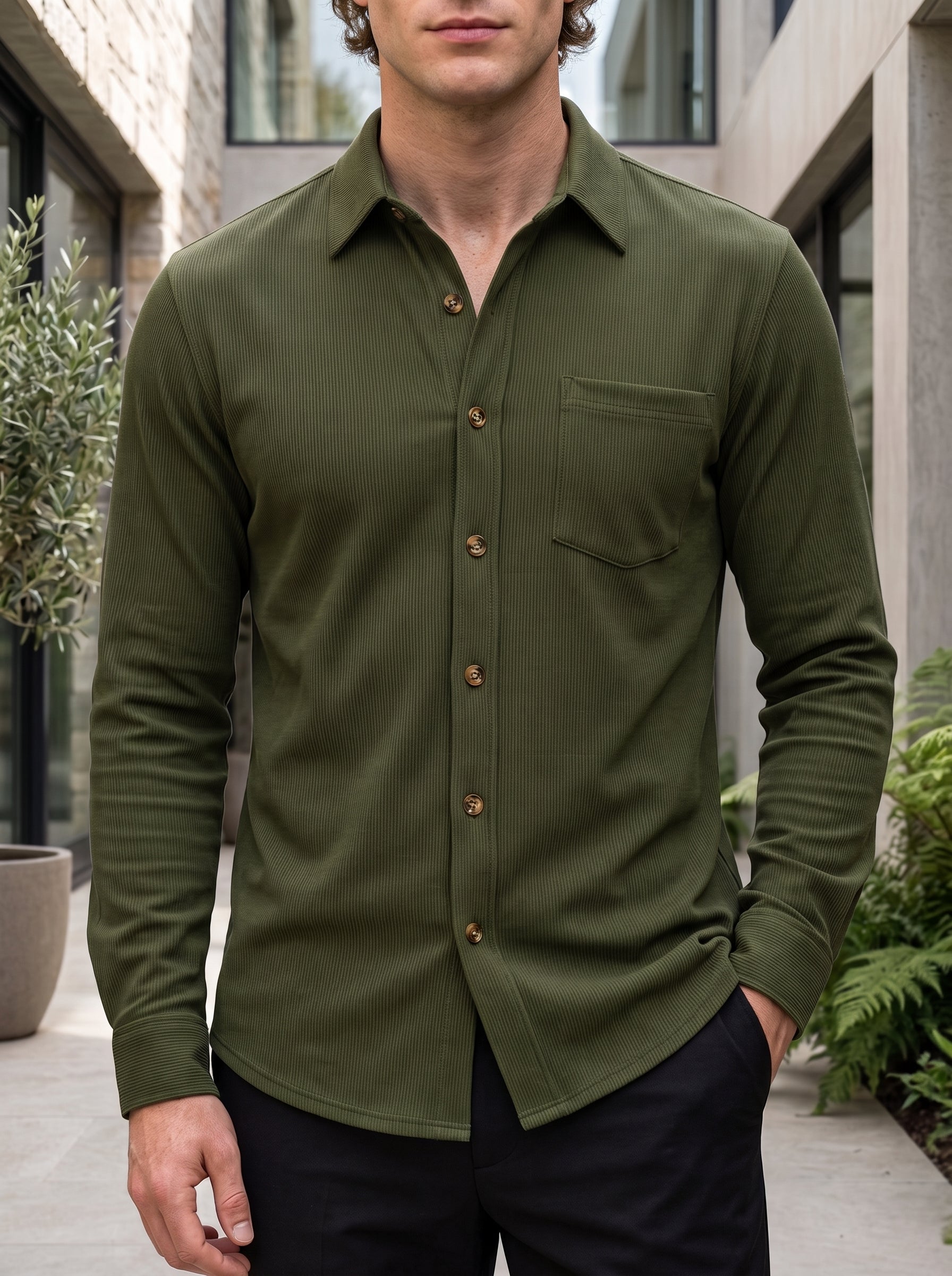 A man wearing an olive green long-sleeve button-down shirt with a chest pocket, paired with dark trousers, standing in an outdoor patio setting.