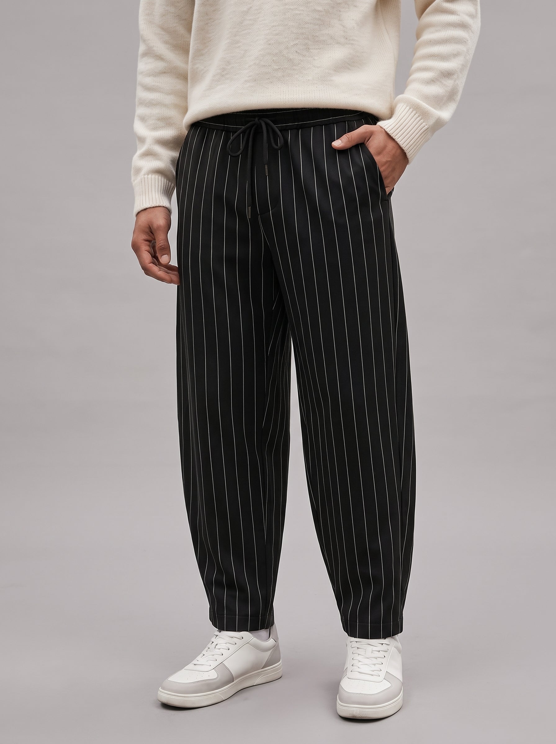 A person wearing black pinstriped jogger-style pants with a drawstring waist, paired with a cream sweater and white sneakers, photographed against a plain light gray studio background.
