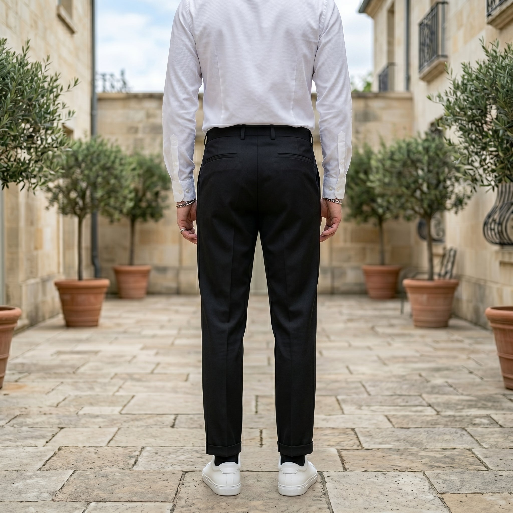 Back view of male model wearing black pleated tailored pants with a white long-sleeve shirt.