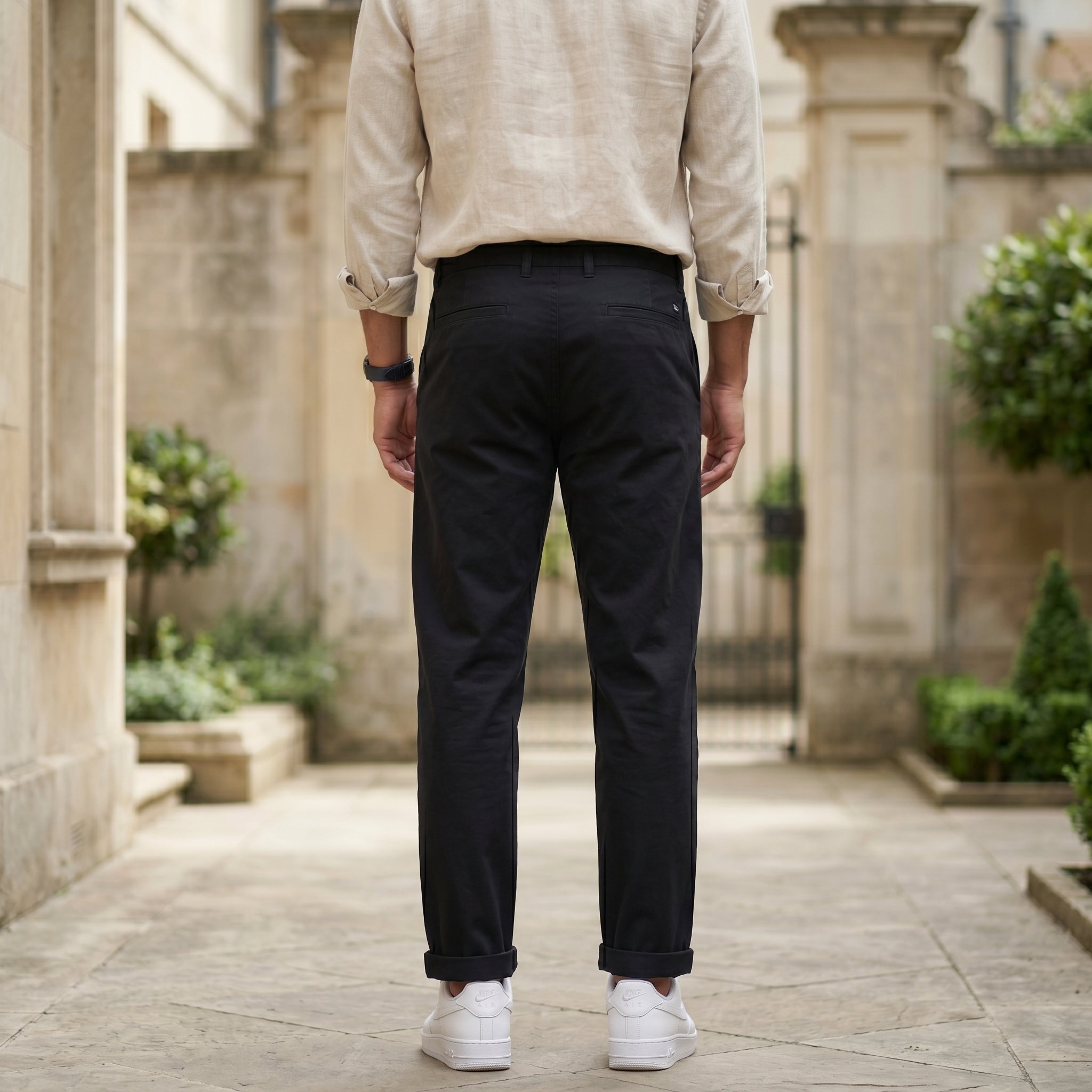 Back view of male model wearing black relaxed-fit casual pants with a beige overshirt and white sneakers.