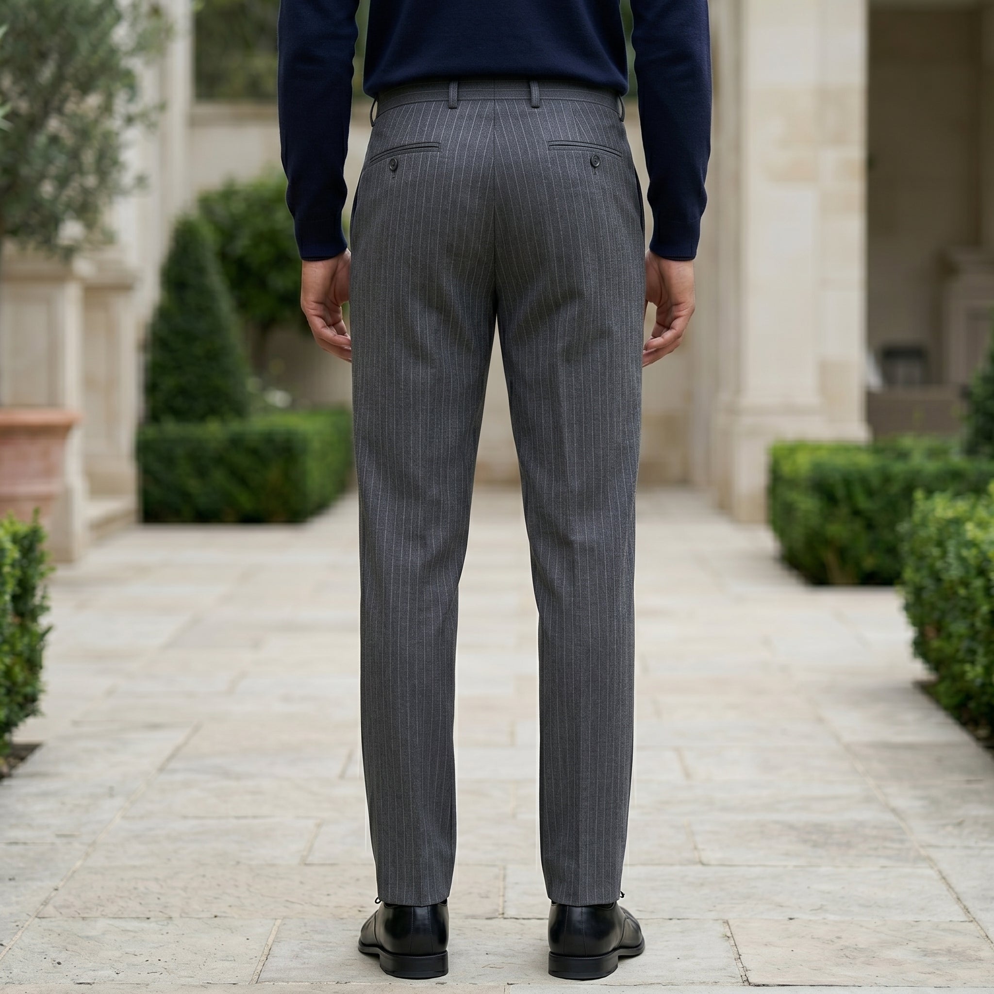 Back view of male model wearing dark gray pleated tailored pants with a navy long-sleeve top.