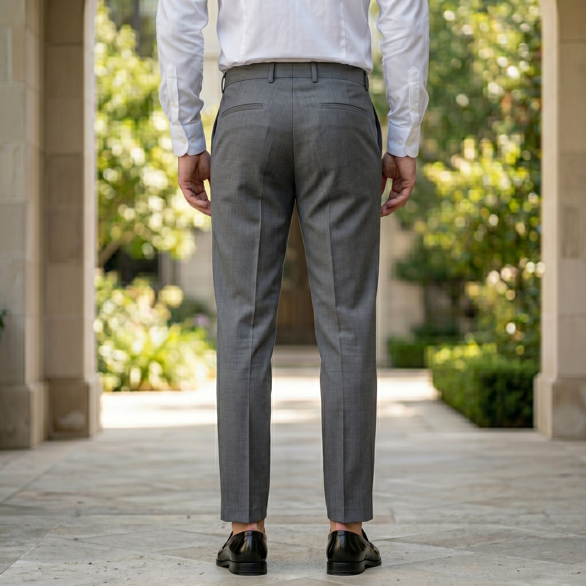 Back view of male model wearing gray pleated tailored pants with a white long-sleeve shirt.