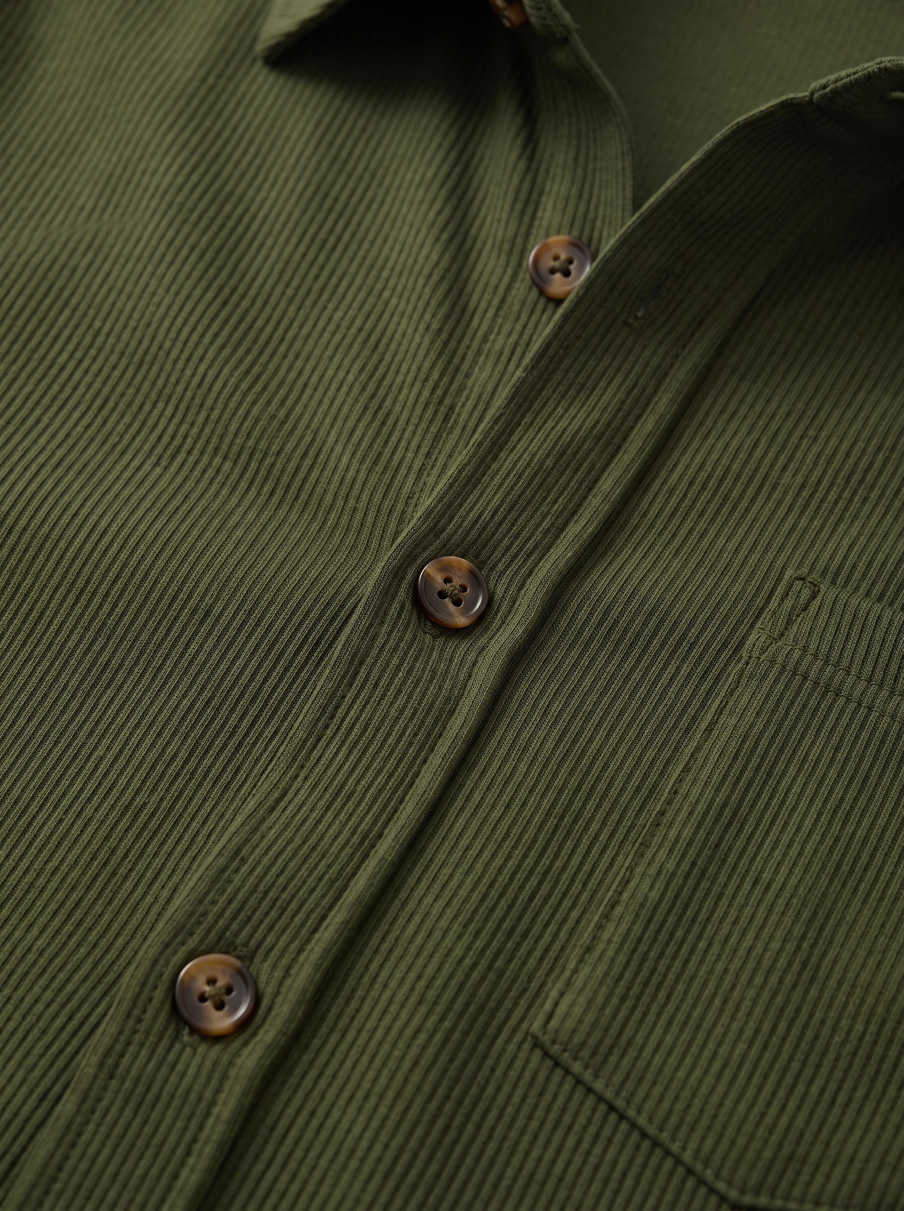 Close-up of an olive green corduroy button-down shirt showing the ribbed texture, brown buttons, and a chest pocket.