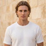 Close-up of male model wearing a white textured short-sleeve summer T-shirt against a beige stone wall.