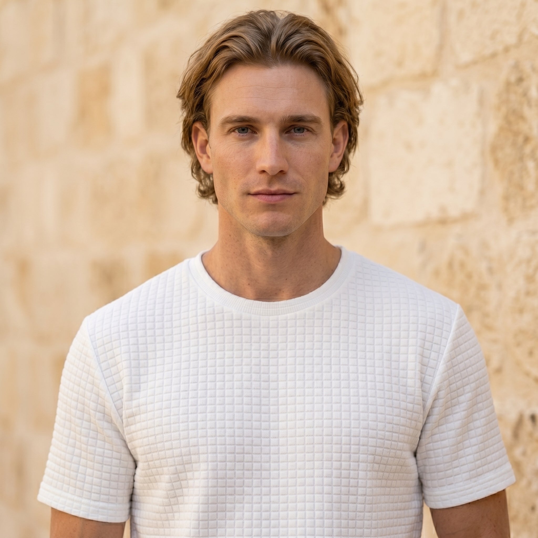 Close-up of male model wearing a white textured short-sleeve summer T-shirt against a beige stone wall.