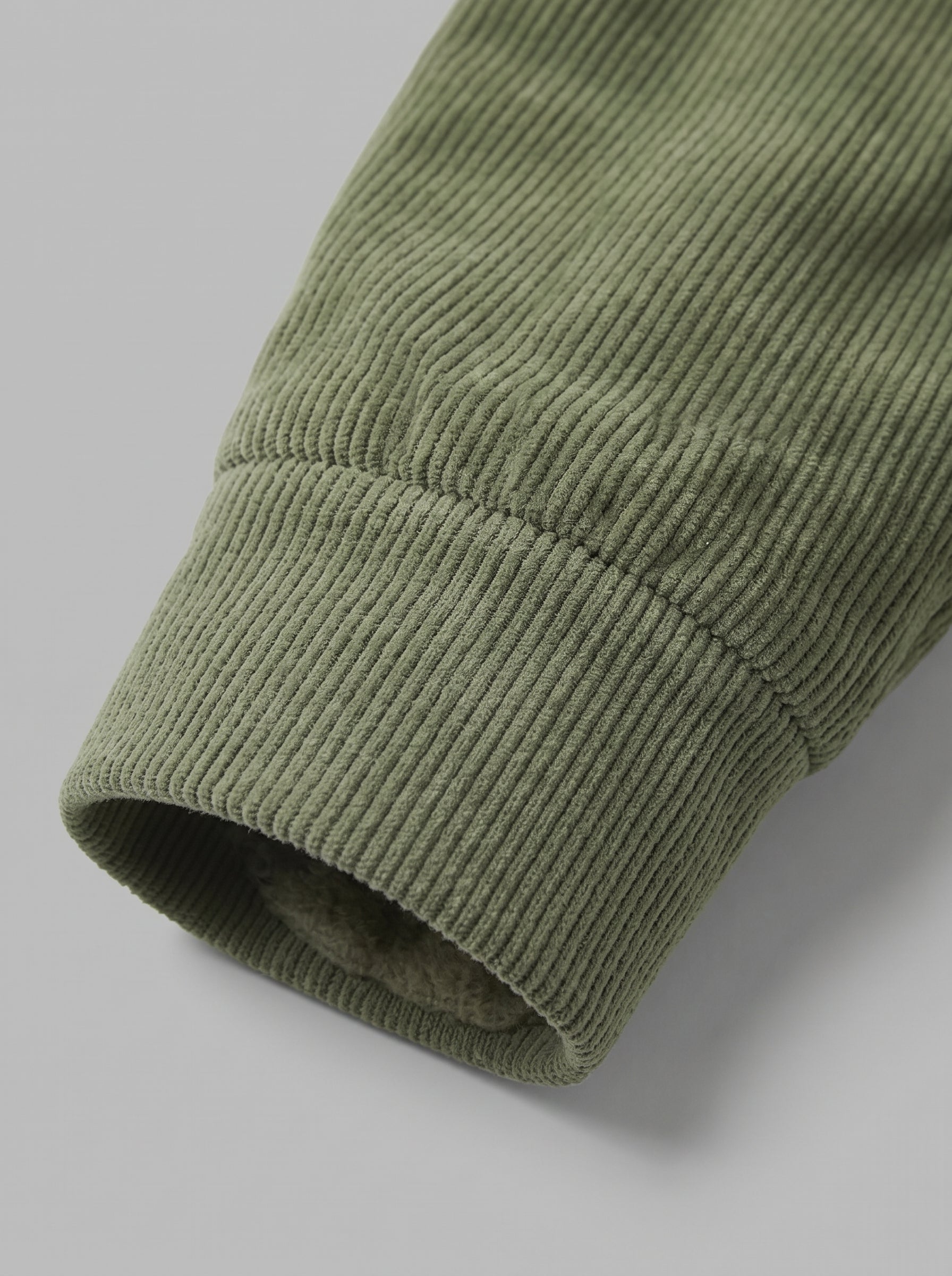 Close-up of a ribbed knit sleeve cuff in muted sage green, showing the fabric texture and a glimpse of a lighter lining inside the opening, set against a plain light gray background.