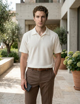 Man in a white polo shirt and brown pants standing outdoors with plants and a building in the background