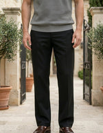 Person wearing a gray sweater and black pants standing outdoors with potted plants and a building in the background.