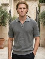 Man wearing a gray patterned polo shirt standing outdoors with stone building and potted plants in the background