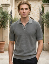 Man wearing a gray patterned polo shirt standing outdoors with stone building and potted plants in the background