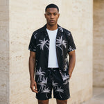 Male model wearing a black palm print short-sleeve shirt over a white T-shirt with matching black drawstring shorts against a beige wall.