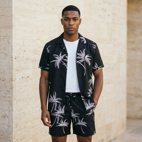 Male model wearing a black palm print short-sleeve shirt over a white T-shirt with matching black drawstring shorts against a beige wall.