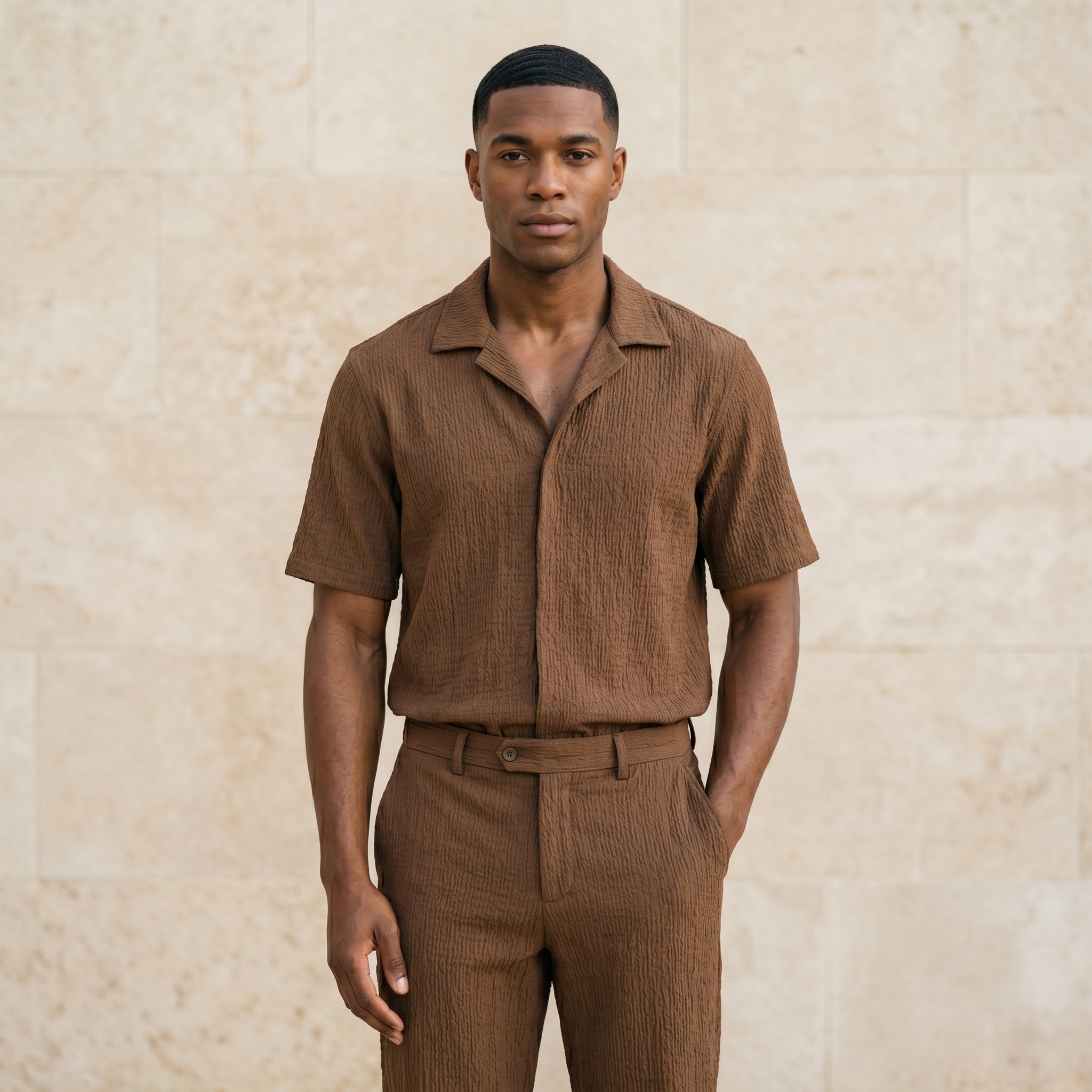 Male model wearing a brown textured short-sleeve shirt and matching pants against a beige stone wall.
