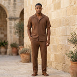 Male model wearing a brown textured short-sleeve shirt with matching straight-leg pants in a stone walkway setting.