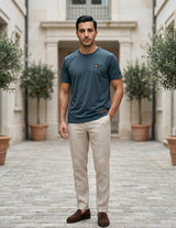 Male model wearing a dark teal blue short-sleeve T-shirt with beige pants in an outdoor walkway setting.