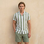 Male model wearing a white and sage green striped short-sleeve shirt with matching sage green drawstring shorts against a beige wall.