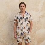 Male model wearing an ivory abstract print short-sleeve button-up shirt with matching summer shorts against a beige wall.