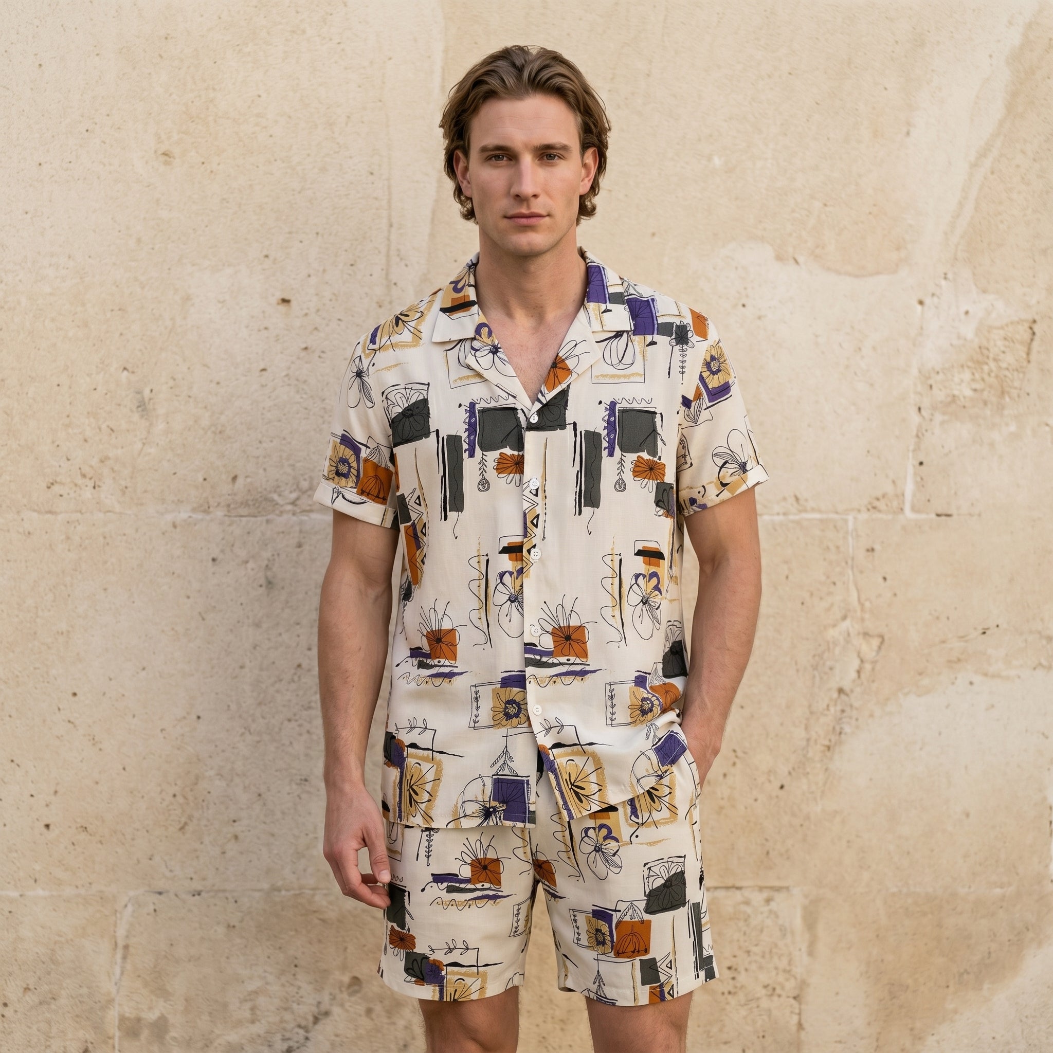 Male model wearing an ivory abstract print short-sleeve button-up shirt with matching summer shorts against a beige wall.