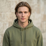 Male model wearing an olive green hooded sweatshirt and matching jogger pants against a beige stone wall.