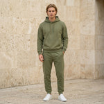 Male model wearing an olive green hoodie with matching jogger pants in a beige stone setting.