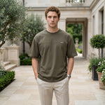 Male model wearing an olive green oversized short-sleeve T-shirt with beige pants in an outdoor walkway setting.