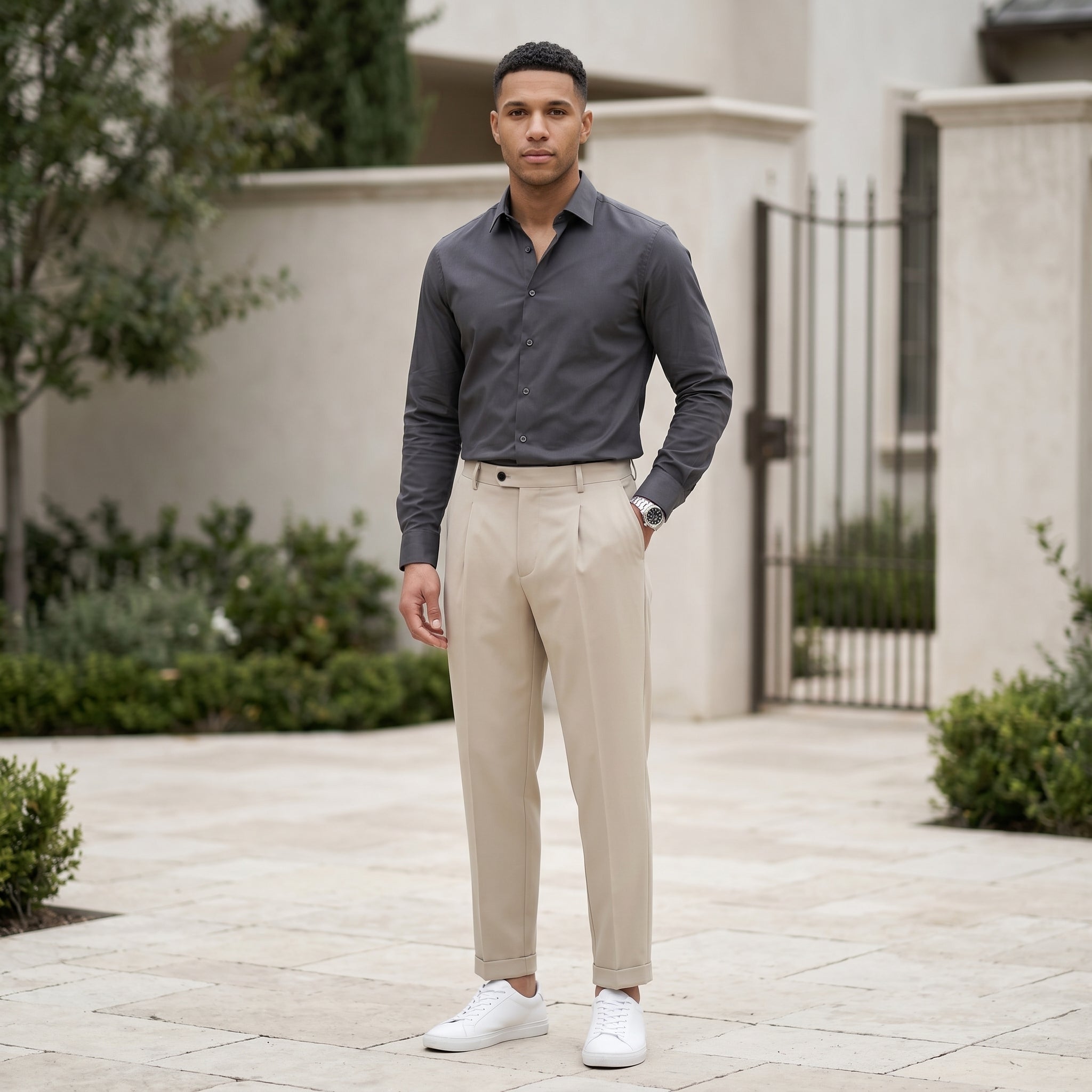 Male model wearing beige pleated casual pants with a dark gray long-sleeve shirt in an outdoor walkway setting.