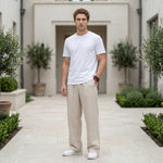 Male model wearing beige textured relaxed-fit pants with a white T-shirt in an outdoor walkway setting.