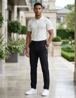 Male model wearing black casual pants with a white short-sleeve polo shirt in an outdoor walkway setting.