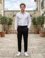 Male model wearing black pleated tailored pants with a white long-sleeve shirt in an outdoor stone walkway setting.